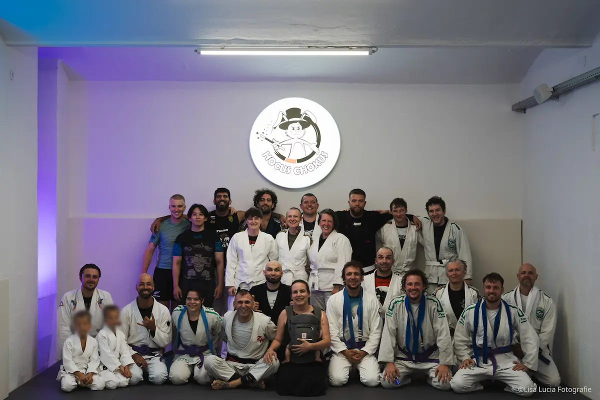 Großes Gruppenfoto der Trainingsgemeinschaft von Hocus Chokus BJJ Wien unter dem leuchtenden Hocus Chokus Logo – Erwachsene und Kinder im Gi und No-Gi, eine familiäre Community