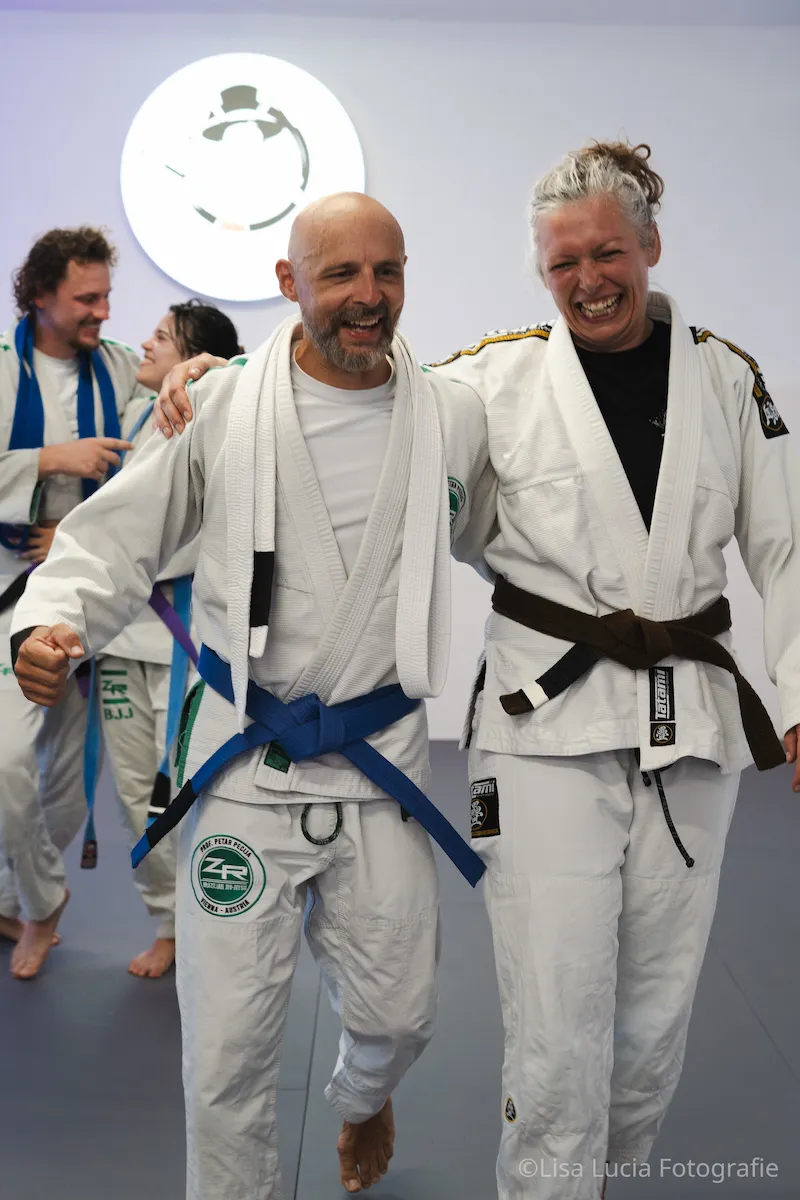 Freudige Gürtelverleihung bei Hocus Chokus BJJ Wien – zwei lachende Trainingspartner im Gi Arm in Arm, frisch beförderter Blaugurt neben Braungurt