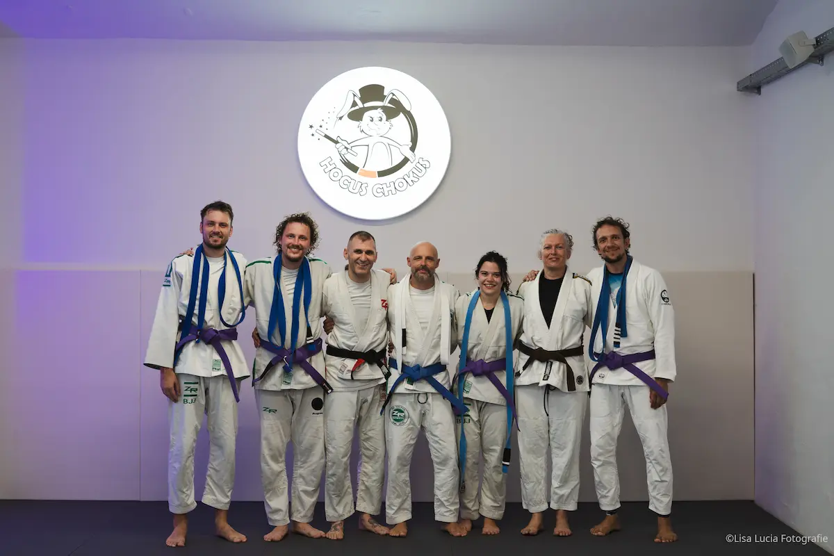 Sieben BJJ-Sportlerinnen und -Sportler im Gi unter dem Hocus Chokus Logo in Wien – Gürtelverleihung mit frisch verliehenen Lila- und Blaugurten