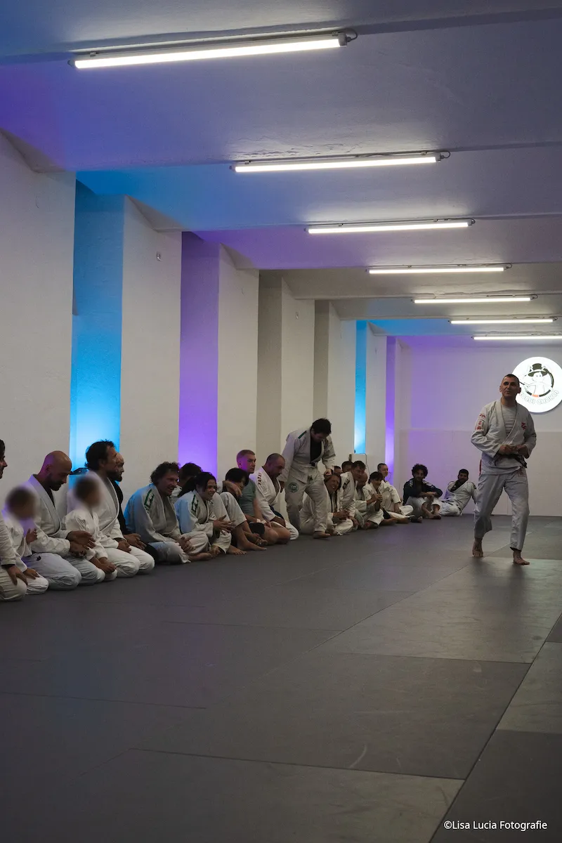 Trainingsgruppe von Hocus Chokus BJJ Wien sitzt aufgereiht am Mattenrand, während der Coach zur Gruppe spricht – stimmungsvolles blau-violettes Licht