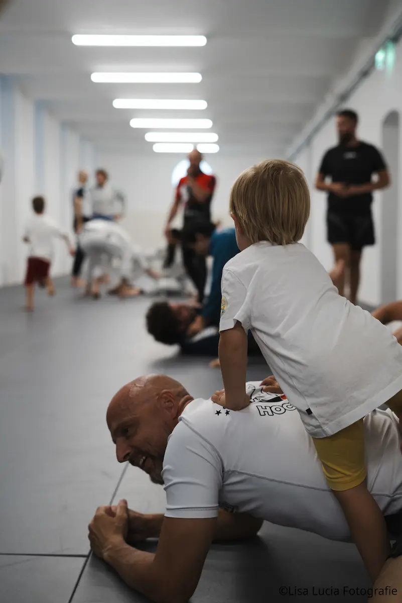 Familienfreundliche Open Mat bei Hocus Chokus BJJ Wien – ein Kind sitzt lachend auf dem Rücken eines Trainierenden, im Hintergrund trainieren Erwachsene und Kinder