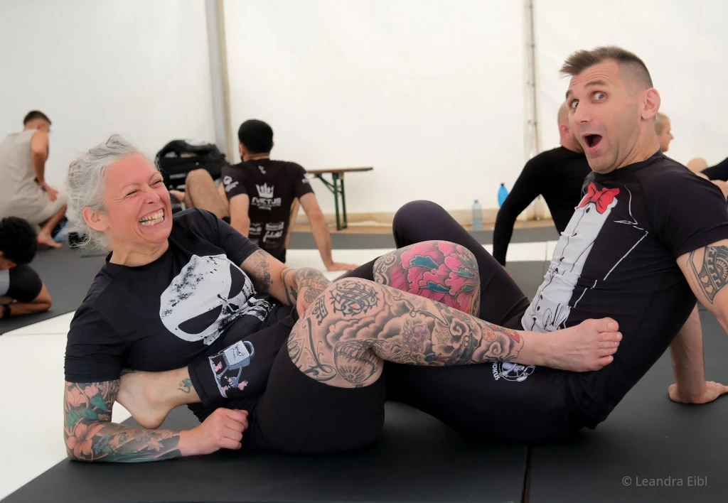 No-Gi Grappling bei Hocus Chokus Wien – Beinverschlingung (Heelhook) im Training, zwei lachende Trainingspartner im Camp-Zelt